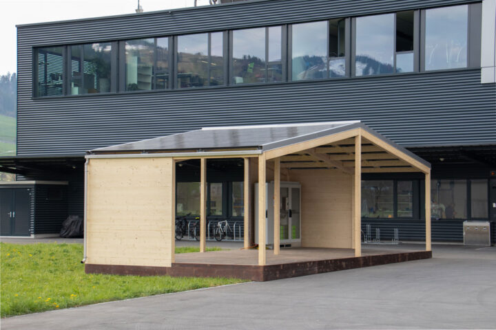 Holzpavillon mit Solarzellen vor einem grauen Gebäude